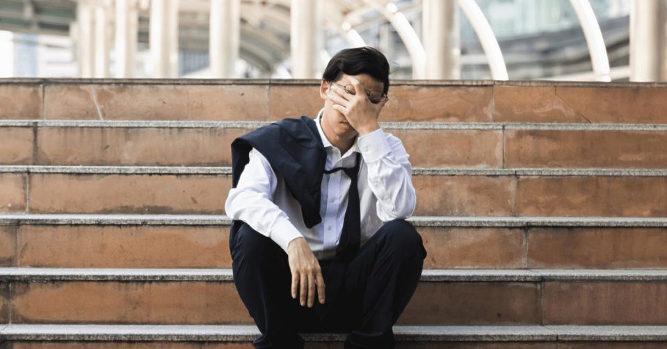 A male office worker sitting on some steps. His head is in one hand and he looks defeated.