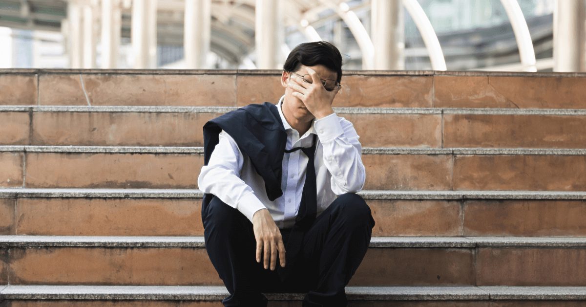 A male office worker sitting on some steps. His head is in one hand and he looks defeated.